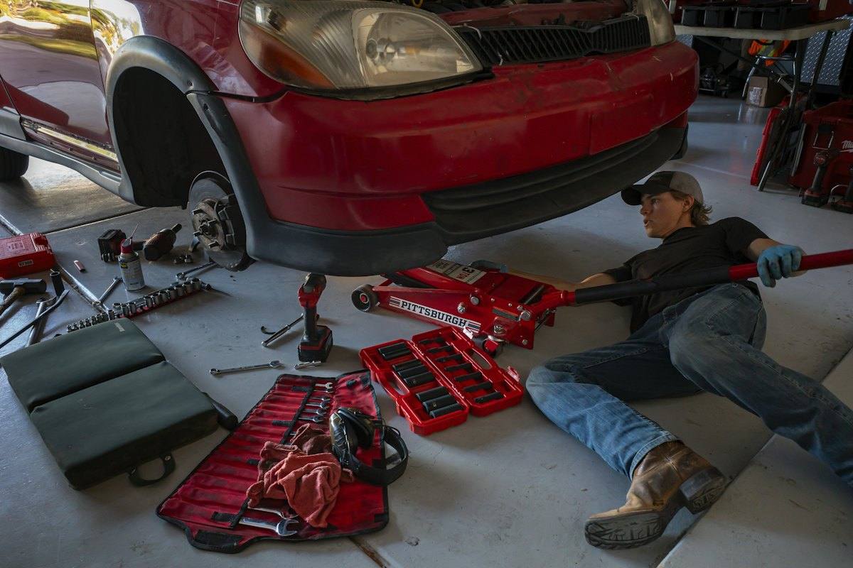 Technician lifting a car with a floor jack beside neatly laid-out tools in the service bay